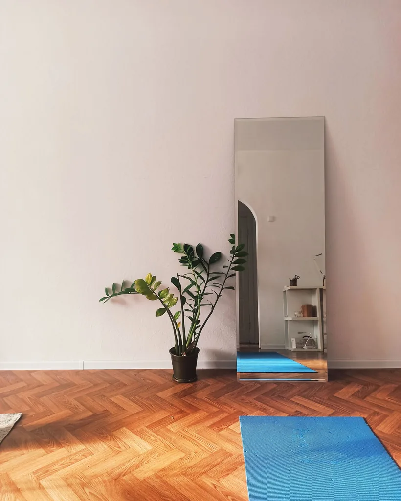 Esterilla de yoga extendida sobre un suelo de madera junto a una planta y un espejo apoyado en la pared